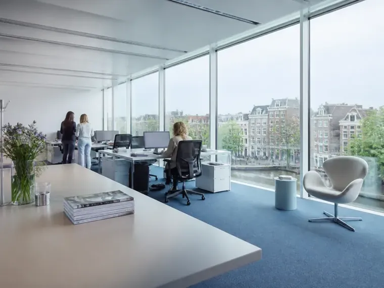 An open office beside a canal, desks along a glass wall and two colleagues talking by the window