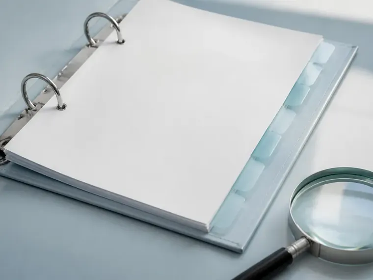 A closed white binder with pale tabbed dividers and a magnifying glass on a desk